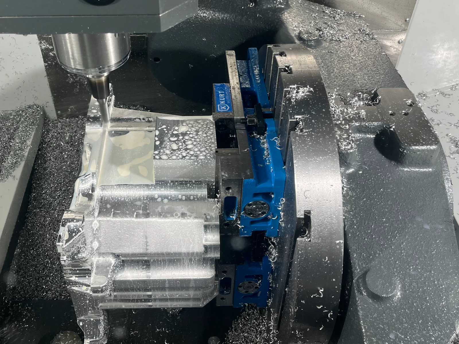 Tool holder and cutter beside a workpiece clamped in a blue vise on a five-axis CNC machine