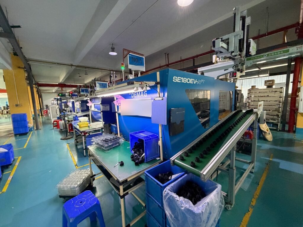 Blue injection molding machine with a robotic arm and a conveyor carrying black molded parts.