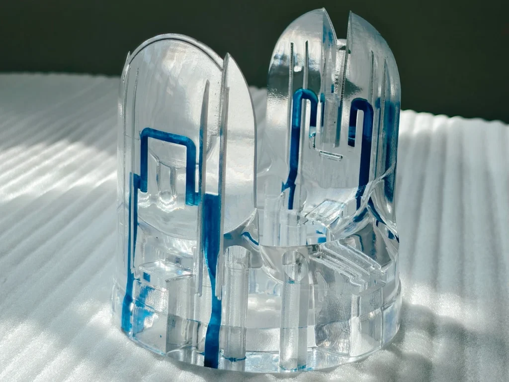 Unpolished transparent resin printed component with blue ink markings indicating internal channel geometry